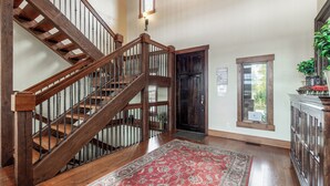 A wooden staircase with a handrail, a red patterned rug, a dark wooden door, and a window with a view of greenery. - Large Home | Reservoir Views & Access | Hot Tub | Fire Pit | Basketball Court (Island Park)