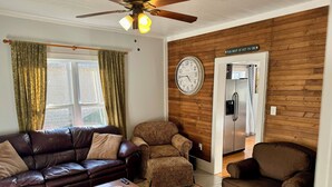 A living room with a brown leather sofa, a patterned armchair, a wooden coffee table, and a red patterned rug. A wall clock and a refrigerator are visible in the background.