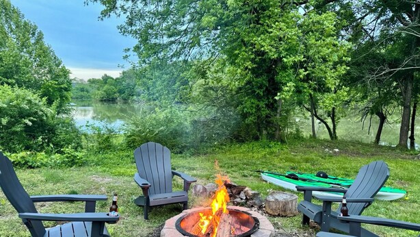 A campfire with two Adirondack chairs and a canoe in a wooded area.