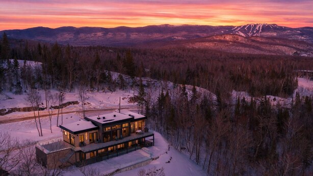 A modern two-story house with a large deck, surrounded by a snowy landscape and dense forest.