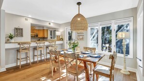 A modern kitchen with wooden bar stools, a dining table set for six, and a large window with a view of trees.