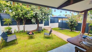 Uma área de pátio com uma mesa posta para refeições, rodeada por cadeiras, e um pequeno jardim com diversas plantas e árvores.