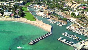 Vista aérea de uma cidade costeira com um porto de iates, praia de areia e edifícios residenciais.