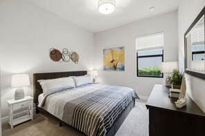 Ein Schlafzimmer mit Bett, Nachttischen, einem Fenster mit Jalousien, einem Wandbild und einer Kommode mit Lampe.