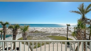 A beach view from a balcony with palm trees, a sandy area, and a clear blue sky.