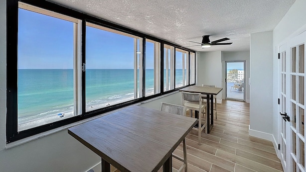A room with large windows overlooking the ocean, a wooden table, and a ceiling fan.