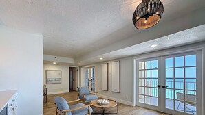 A spacious living room with a wooden floor, a round coffee table, two blue armchairs, and a large window with a view of the sea.