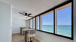A room with a large window overlooking the ocean, a ceiling fan, and a dining area with wooden furniture.