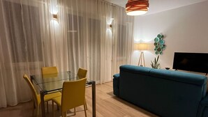 Moderni olohuone, jossa on lasinen ruokapöytä, keltaiset tuolit, turkoosi sohva, televisio ja lamppu.