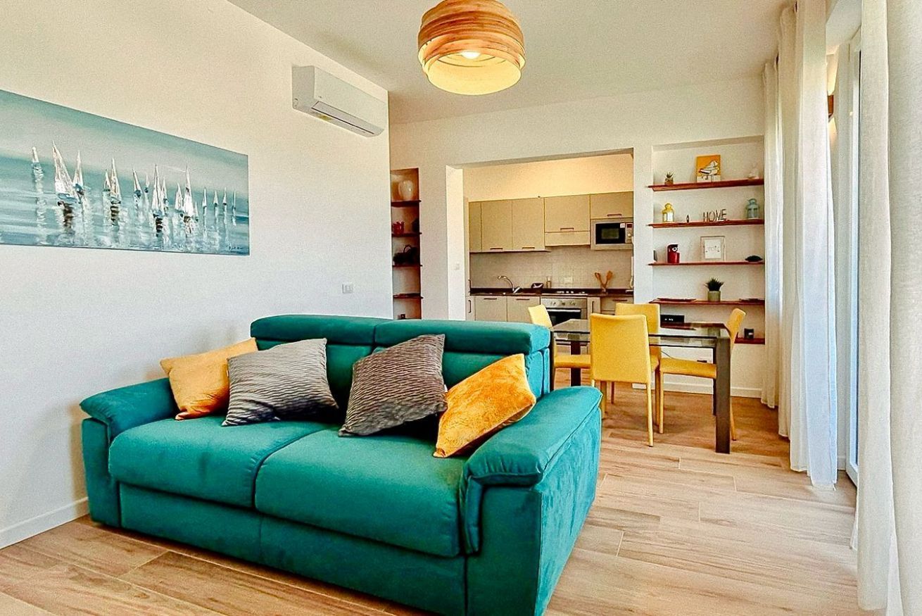 Ein blaugrünes Sofa mit Kissen, ein Esstisch mit Stühlen, ein Küchenbereich und ein Gemälde an der Wand.