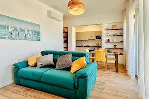 A teal sofa with cushions, a dining table with chairs, a kitchen area, and a painting on the wall. - SE127 - Senigallia, two-room apartment with amenities and air conditioning (Senigallia)