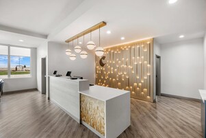 Un área de recepción moderna con un mostrador blanco, iluminación colgante y una pared de madera con paneles iluminados.