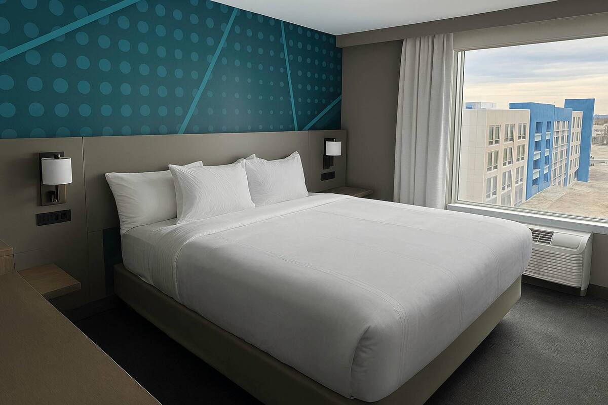 A hotel room with a large bed, two bedside lamps, a window with a view of buildings, and a wall with a blue polka dot pattern.