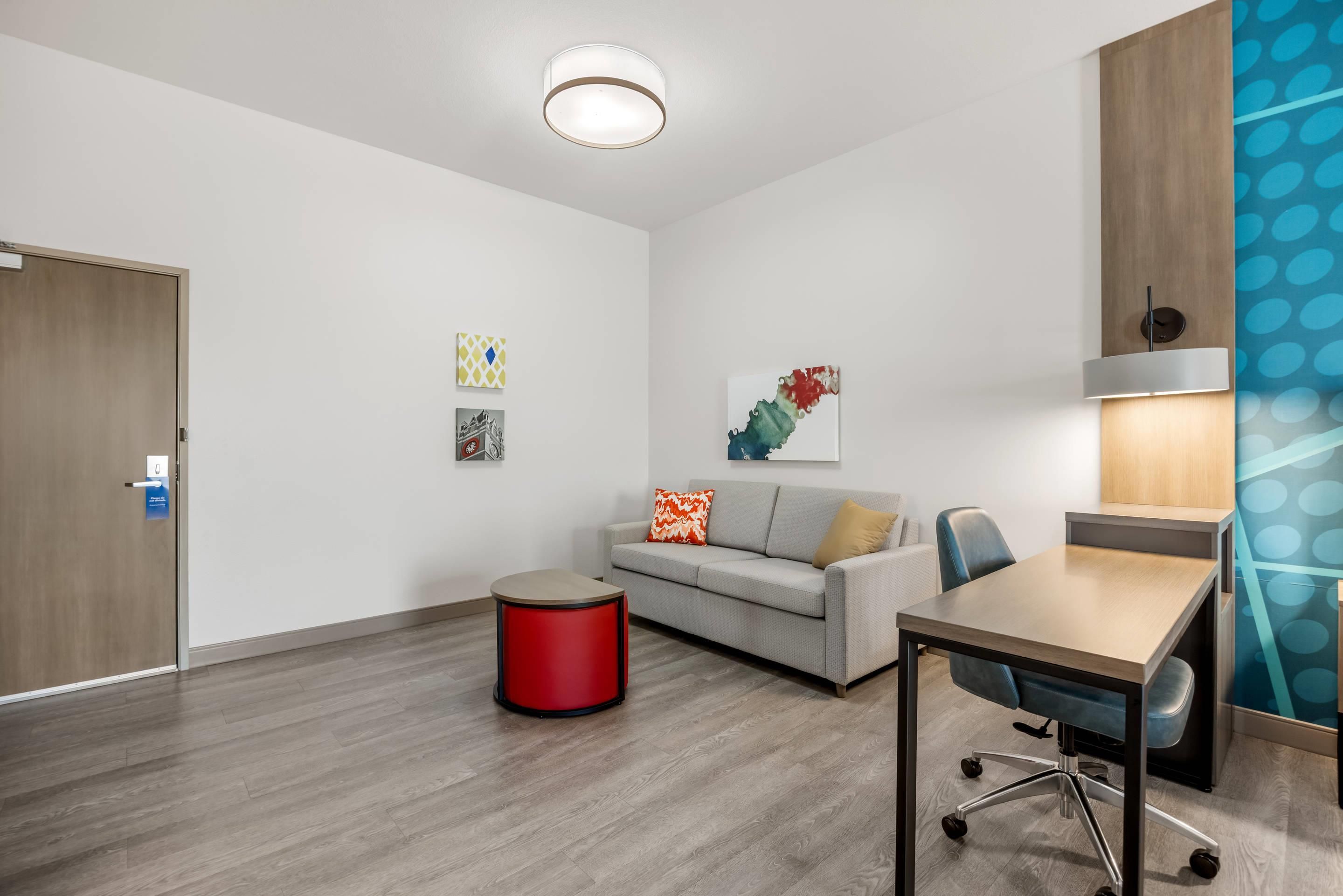 A modern hotel room with a wooden floor, a grey sofa, a red accent chair, a wooden desk, and a lamp.