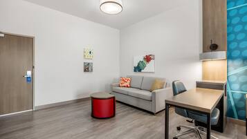 A modern hotel room with a wooden floor, a grey sofa, a red accent chair, a wooden desk, and a lamp.