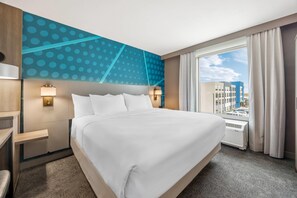 A hotel room with a large bed, a desk, a chair, a window with curtains, and a view of buildings outside. - Comfort Inn & Suites Colby (Colby)
