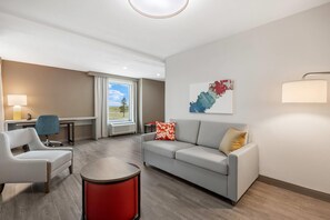 A modern hotel room with a sofa, armchair, and a small table. There is a desk with a lamp, a window with curtains, and a painting on the wall. - Comfort Inn & Suites Colby (Colby)