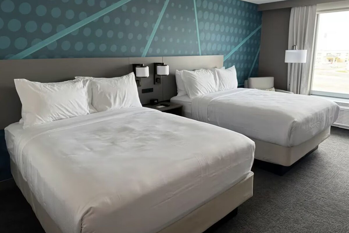 Una habitación de hotel con dos camas, cabeceros, lámparas de noche, una ventana con cortinas y una pared con un diseño geométrico.