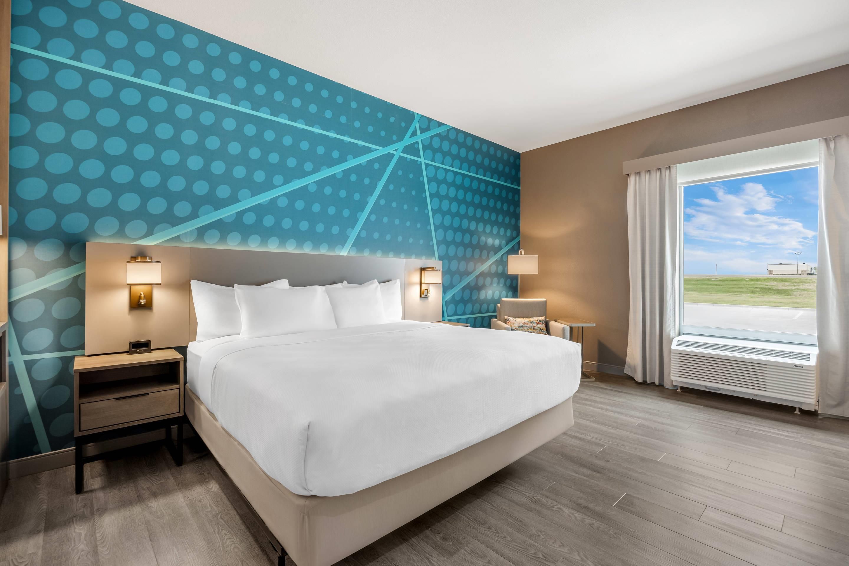 A hotel room with a large bed, two bedside tables with lamps, a view of the outdoors through a window, and a wall with a blue polka dot pattern.