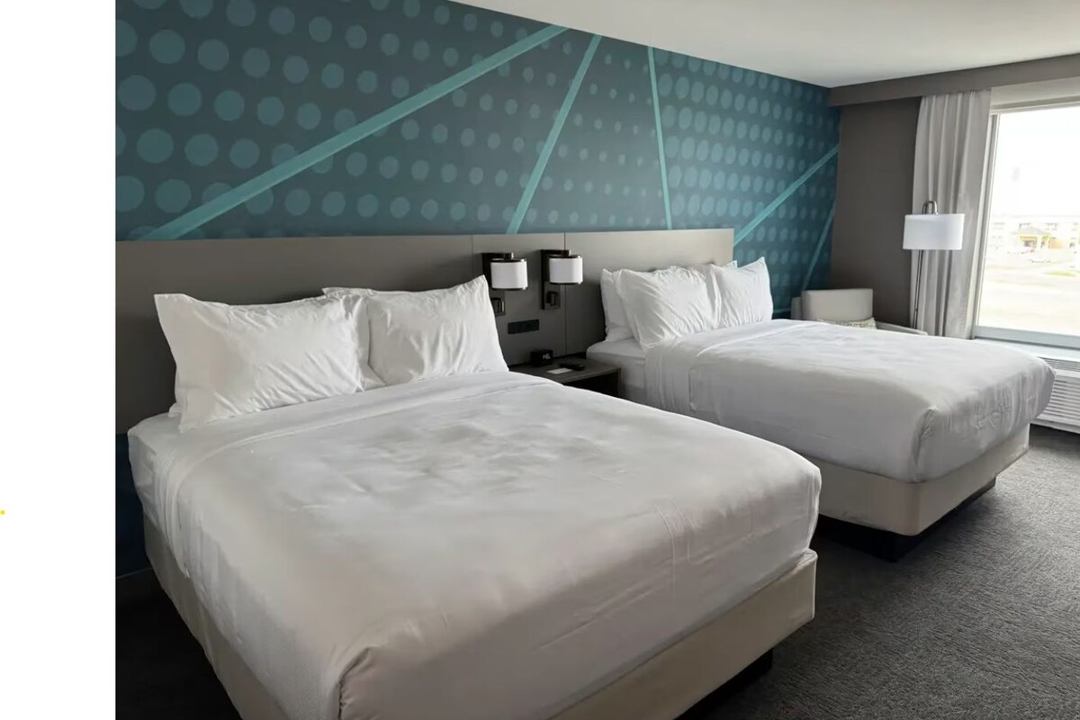Habitación de hotel con dos camas, cabecera con luces empotradas, mesita de noche y ventana con cortinas.