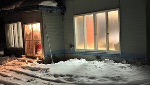 A building with a snow-covered ground, a visible door, and a window with light inside.