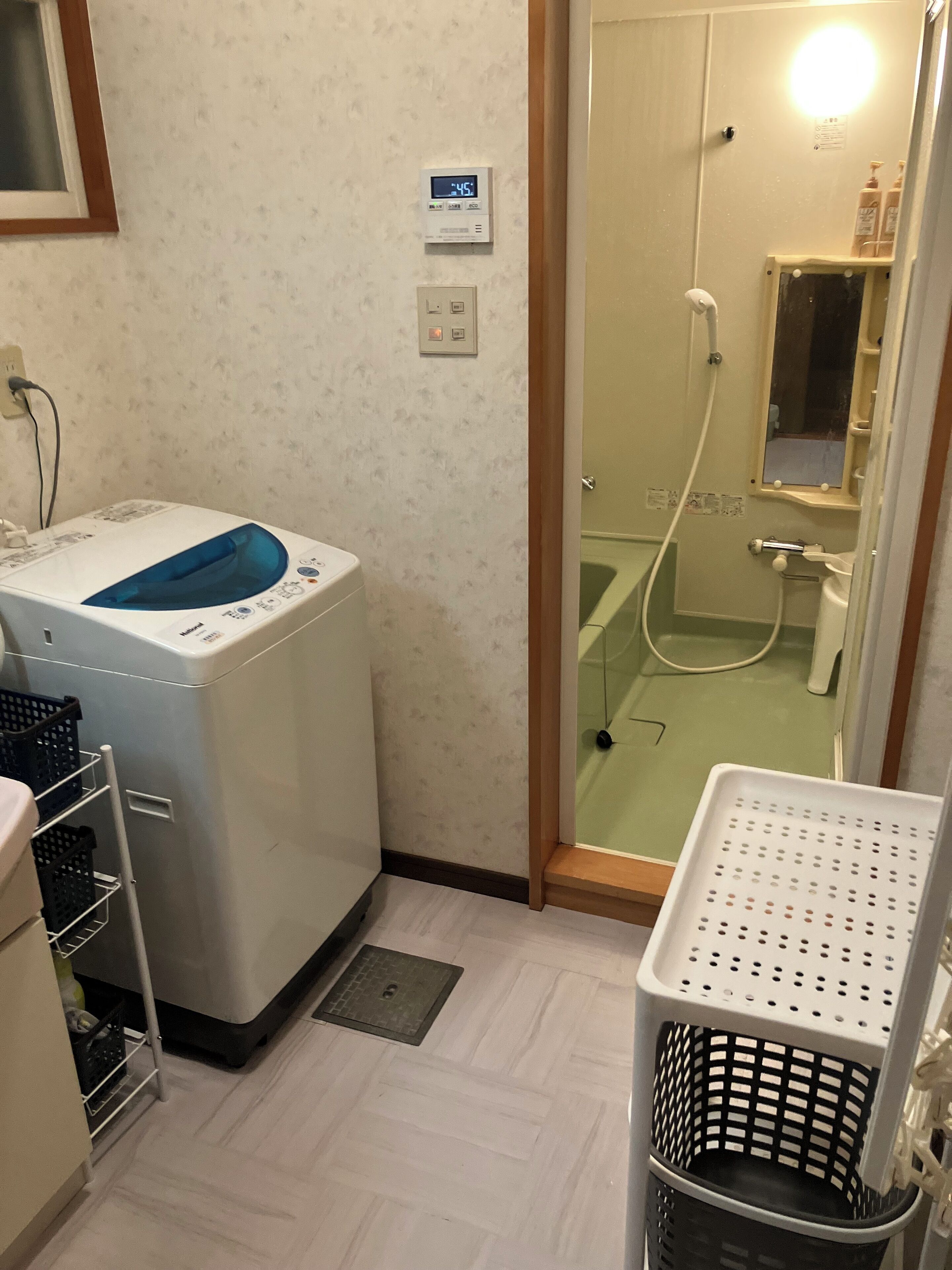 A laundry area with a washing machine, a dryer, a utility shelf, and a glass-enclosed shower area with a mirror and toiletries.