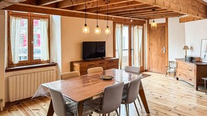Une table à manger en bois, des chaises, un téléviseur et un meuble dans une pièce dotée de poutres en bois et de grandes fenêtres.