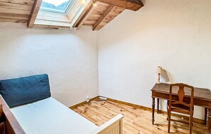 A room with a sloped ceiling, a bed with a blue pillow, a wooden desk with a lamp, and a chair.