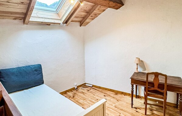 Una habitación con techo inclinado, una cama con almohada azul, un escritorio de madera con lámpara y una silla.