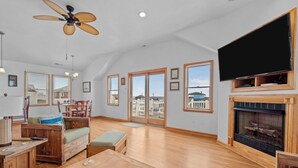 A spacious living room with a fireplace, wooden furniture, large windows, and a ceiling fan. - 4744- Sandy Beaches, a spacious home in the heart of Kill Devil Hills! (Kitty Hawk)