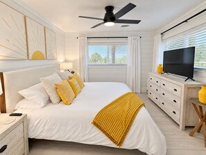 A bedroom with a bed, bedside tables, a TV, a ceiling fan, and a window with blinds.