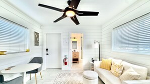 Une pièce moderne et bien éclairée, dotée d’un ventilateur de plafond, d’un canapé blanc agrémenté de coussins à motifs, d’un pouf rond et d’un coin repas avec une table et des chaises blanches.