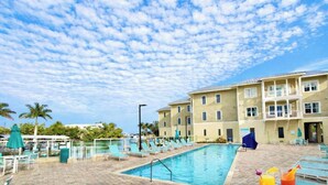 Une piscine avec des chaises longues et des parasols, un bâtiment doté de balcons et un ciel dégagé.