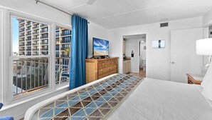 A hotel room with a large bed, a view of the city through a window, a ceiling fan, and a wooden dresser. - Experience Beachfront Luxury w/ Spectacular Views! Pool & Hot Tub (South Padre Island)