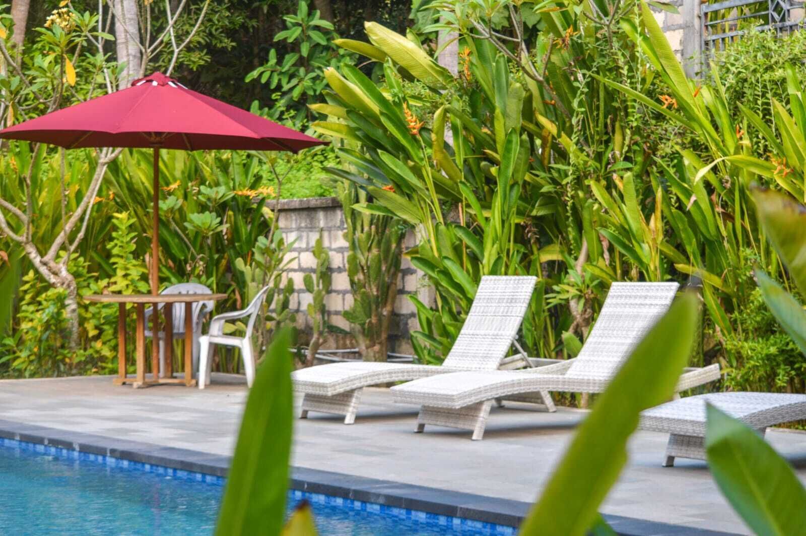 Zona de piscina con sillones de descanso, una sombrilla roja y abundante vegetación.