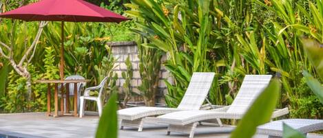 Zona de piscina con sillones de descanso, una sombrilla roja y abundante vegetación.