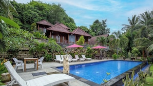 A swimming pool with lounge chairs and umbrellas, surrounded by lush greenery and traditional-style buildings. - Coconut Hill Cottage (Penida Island)