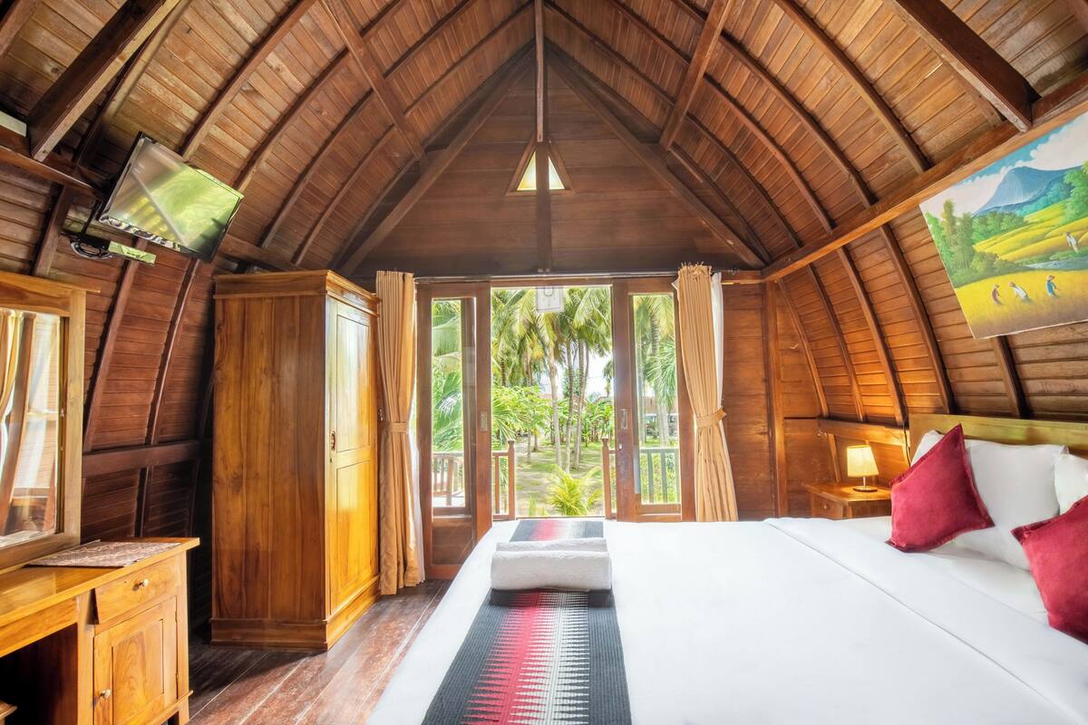 A room with a wooden ceiling, a bed with white and red pillows, a wooden desk, and a window with curtains.