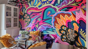 A vibrant room with a colorful abstract mural, a glass cabinet, a table with a plant, and a chair with a fur throw.