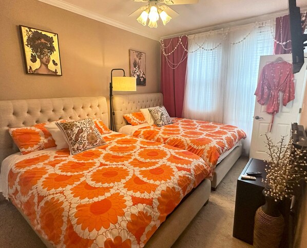 Un dormitorio con cabecera capitonada, dos camas con ropa de cama floral naranja, una lámpara de pie y obras de arte en las paredes.