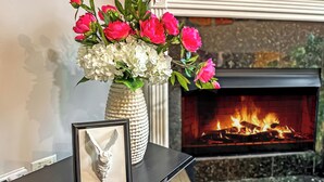 A fireplace with a fire, a vase with pink and white flowers, and a framed picture on a black table.