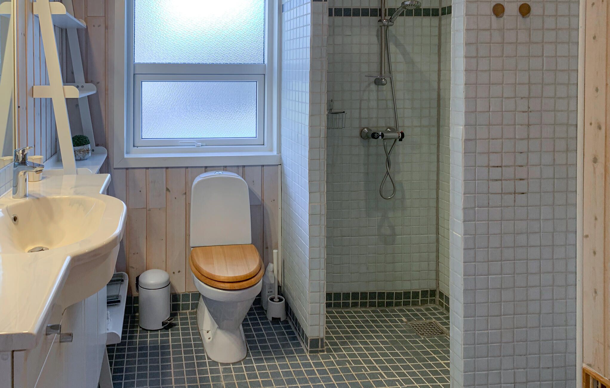 A bathroom with a wooden toilet, a white sink, a shower with a glass door, and a window above the toilet.
