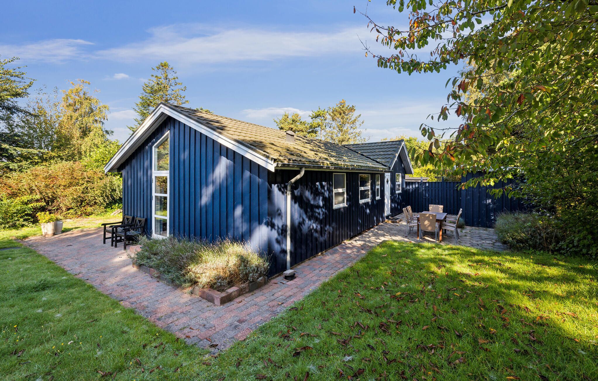 A blue wooden cabin with a brick patio and a small deck, surrounded by a garden and a wooden fence.
