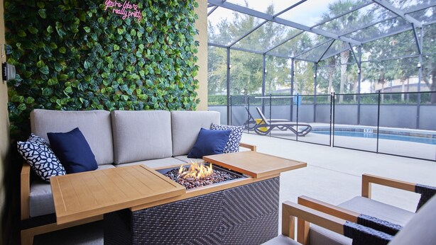 A modern outdoor seating area with a fire feature, a sofa with patterned cushions, and a green leafy wall. - Luxury Disney Getaway with Pool (Davenport)