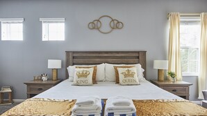 A neatly made bed with a white and gold bedspread, two pillows with "King" and "Queen" labels, a wooden headboard, bedside tables with lamps, and a wall art piece above the bed. - Luxury Disney Getaway with Pool (Davenport)