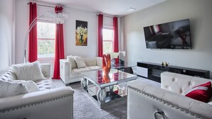 A modern living room with a glass coffee table, white sofas, red curtains, and a flat-screen TV on the wall. - Luxury Disney Getaway with Pool (Davenport)