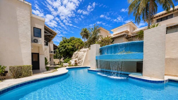 A swimming pool with a waterfall feature, surrounded by buildings with a light-colored exterior and palm trees. - 4BR House | Hotel Zone | Beach & 3 Pools (Cancún)