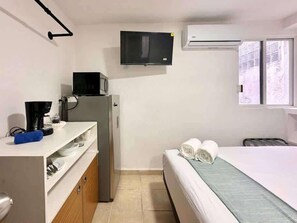 A compact hotel room with a bed, a small kitchenette, a TV, and an air conditioning unit.