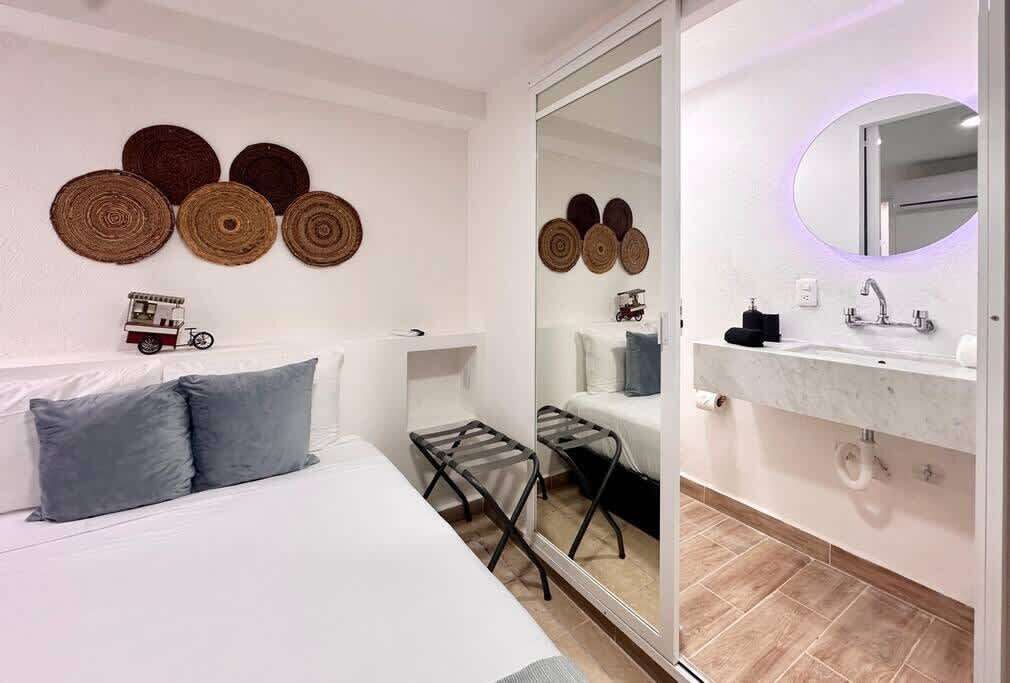 A modern hotel room with a bed, a small stool, a mirror, and a bathroom with a sink and toilet.