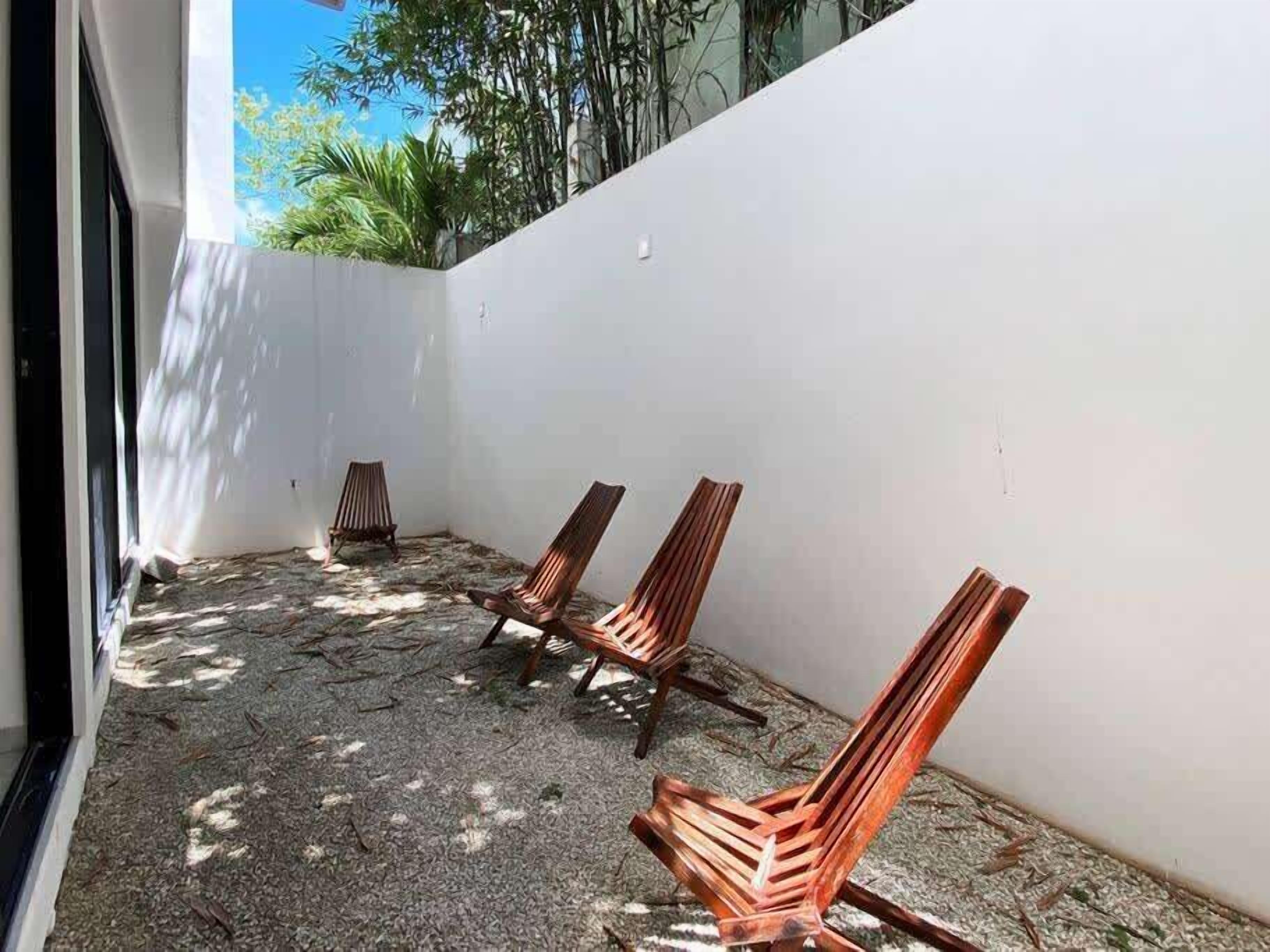A covered outdoor seating area with three wooden chairs and a white wall.